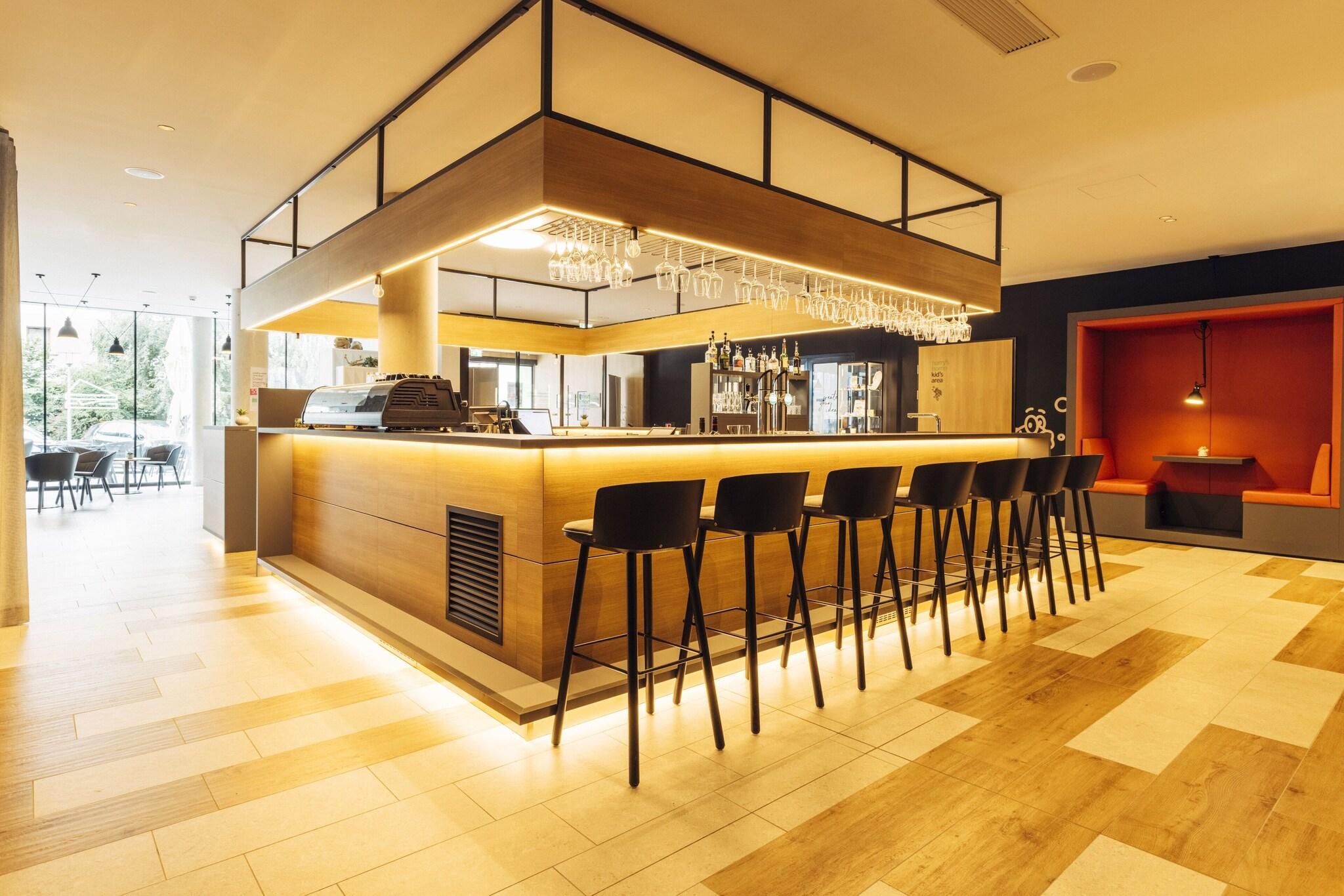 Uma área de bar moderna com balcão de madeira, banquetas pretas e um candelabro.