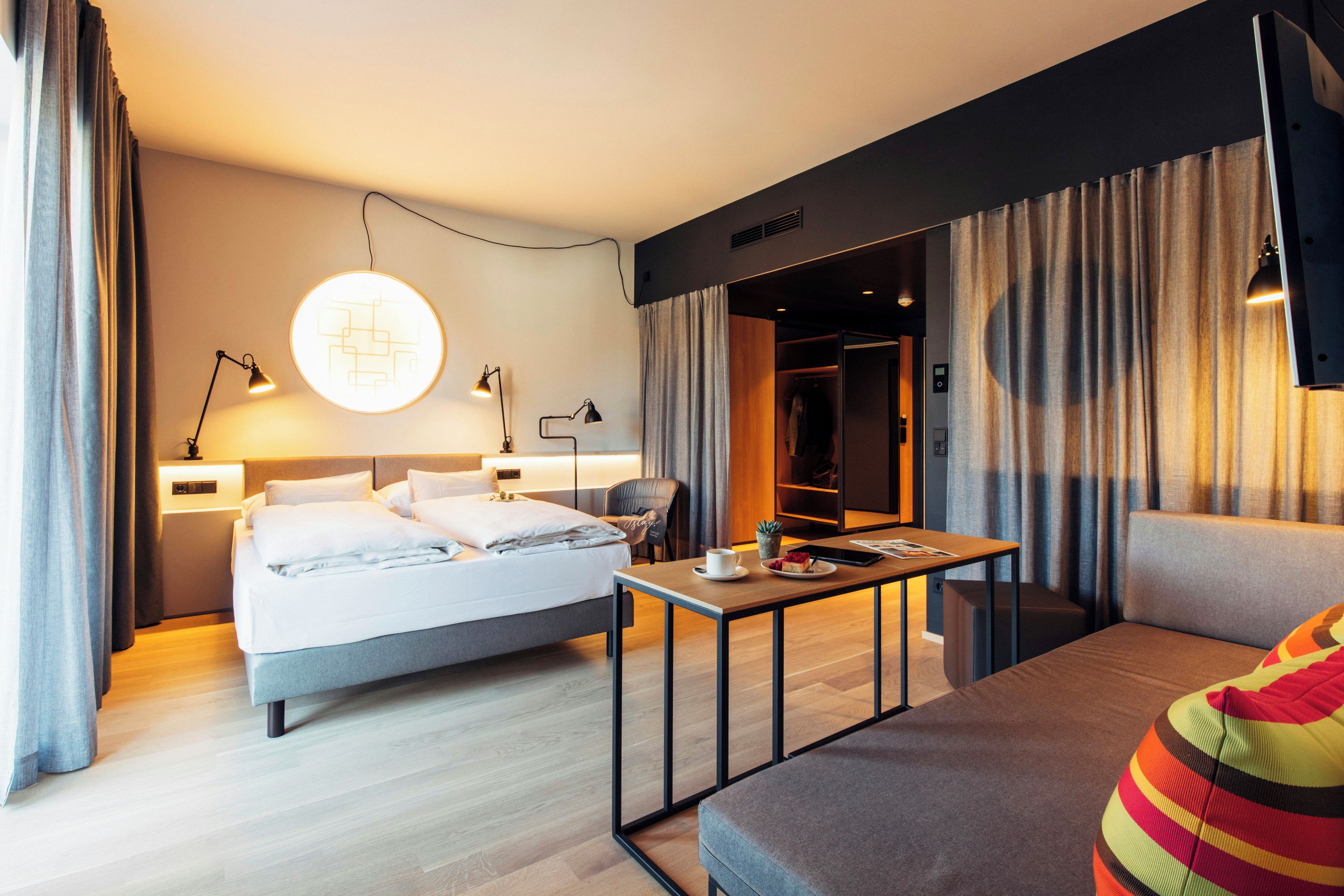 Una habitación de hotel moderna con cama, mesitas de noche, escritorio con silla, sofá y un ventanal con cortinas.