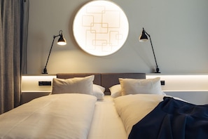 Quarto de hotel com design moderno, com uma cama grande com roupa de cama branca, duas mesas de cabeceira, um ornamento redondo na parede e duas luminárias fixadas na parede.
