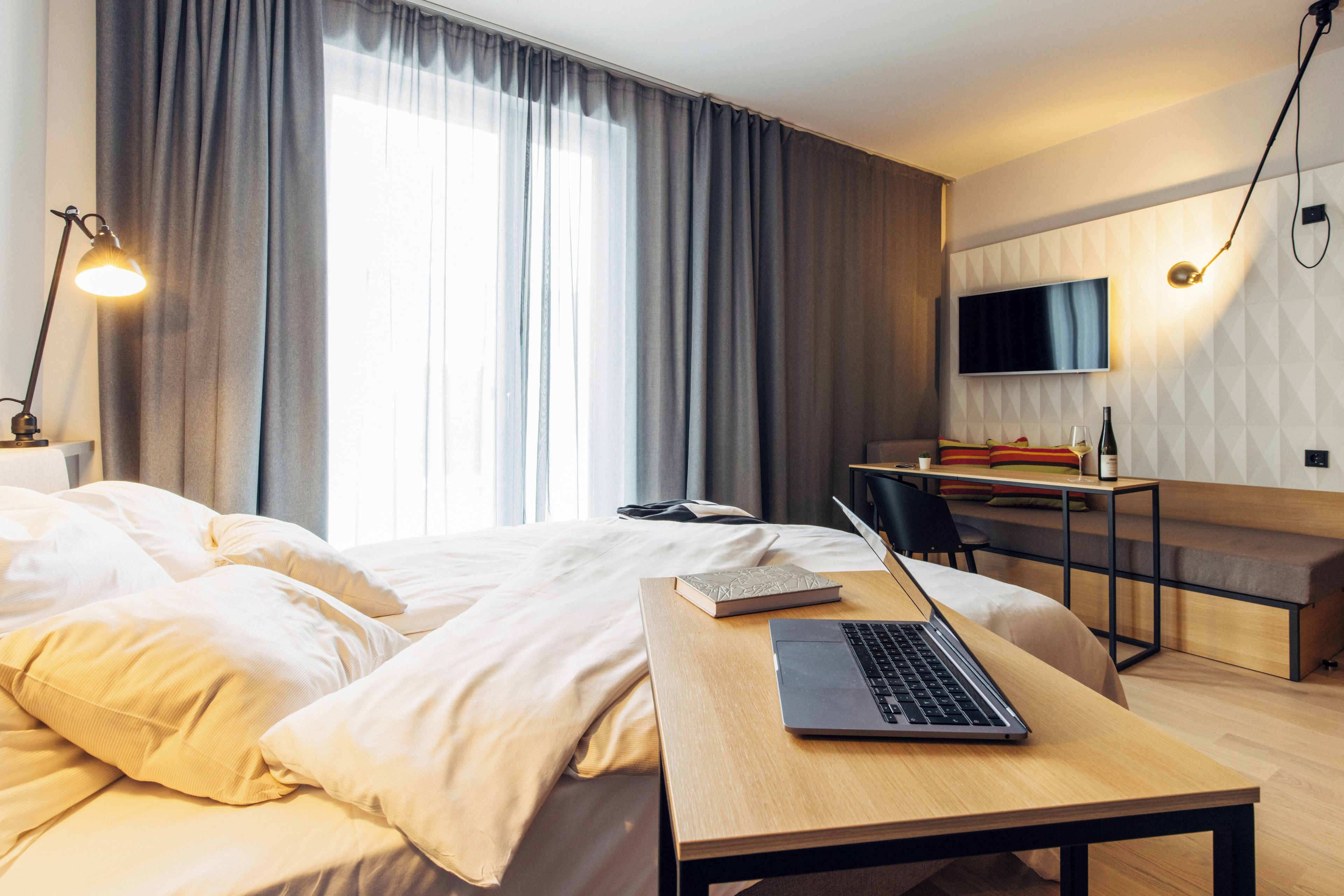 A hotel room with a bed, a desk with a laptop, a chair, a TV, and a window with curtains.