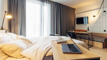 A hotel room with a bed, a desk with a laptop, a chair, a TV, and a window with curtains.