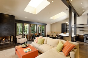 A modern living room with a fireplace, large windows, a dining area, and a kitchen.