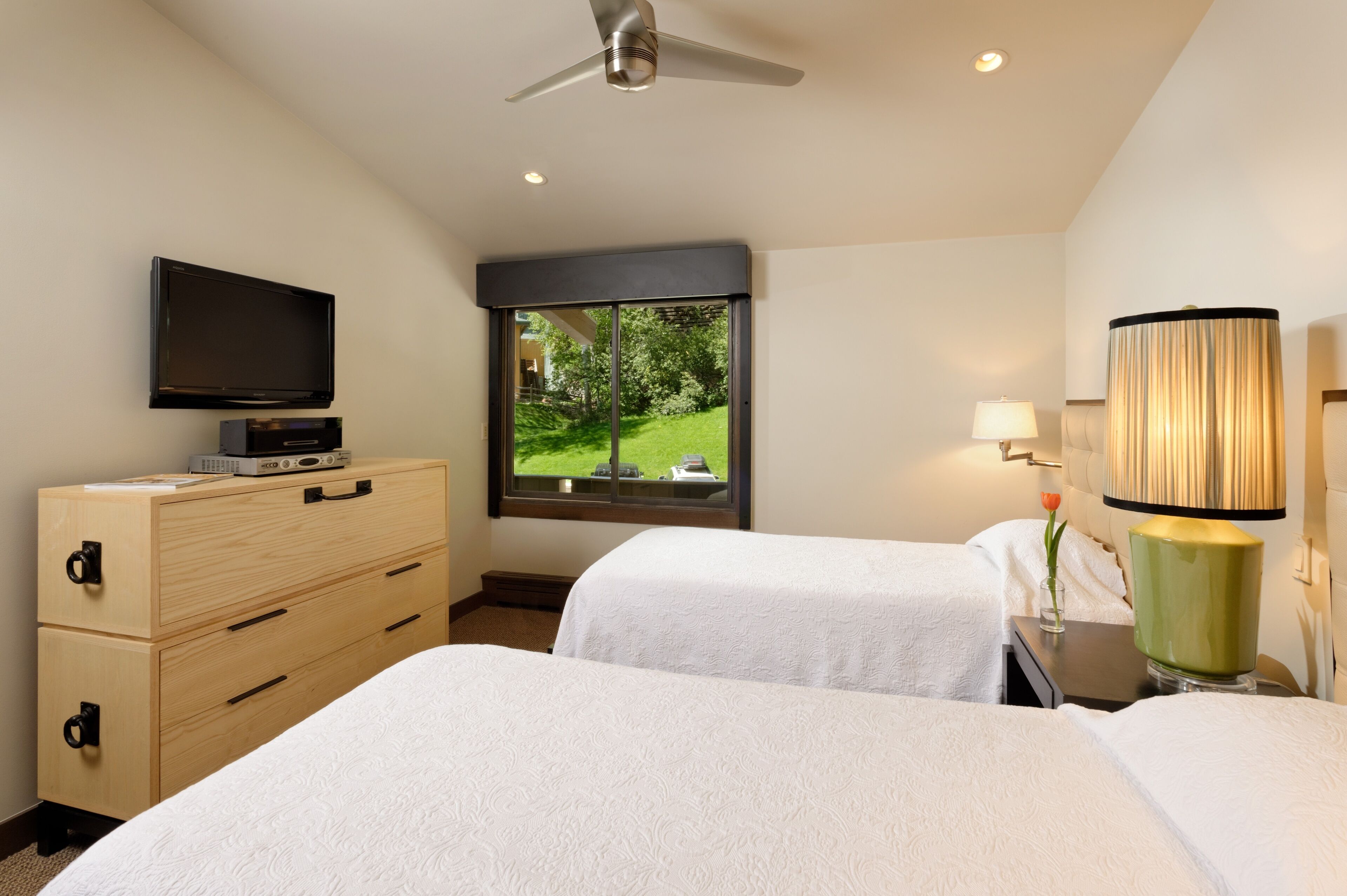 Quarto de hotel com duas camas, uma televisão, penteadeira, janela vista para área verde e abajur.