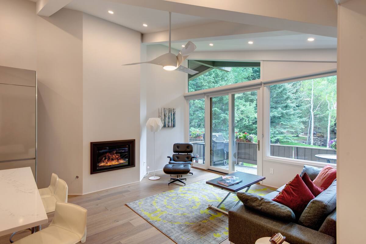 A modern living room with a fireplace, a ceiling fan, and large windows offering a view of the outdoors.