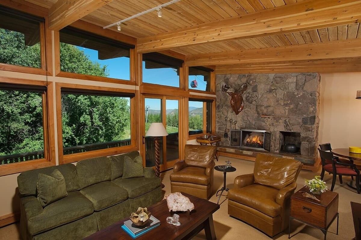 A cozy living room with a stone fireplace, leather furniture, and large windows offering a view of the outdoors.