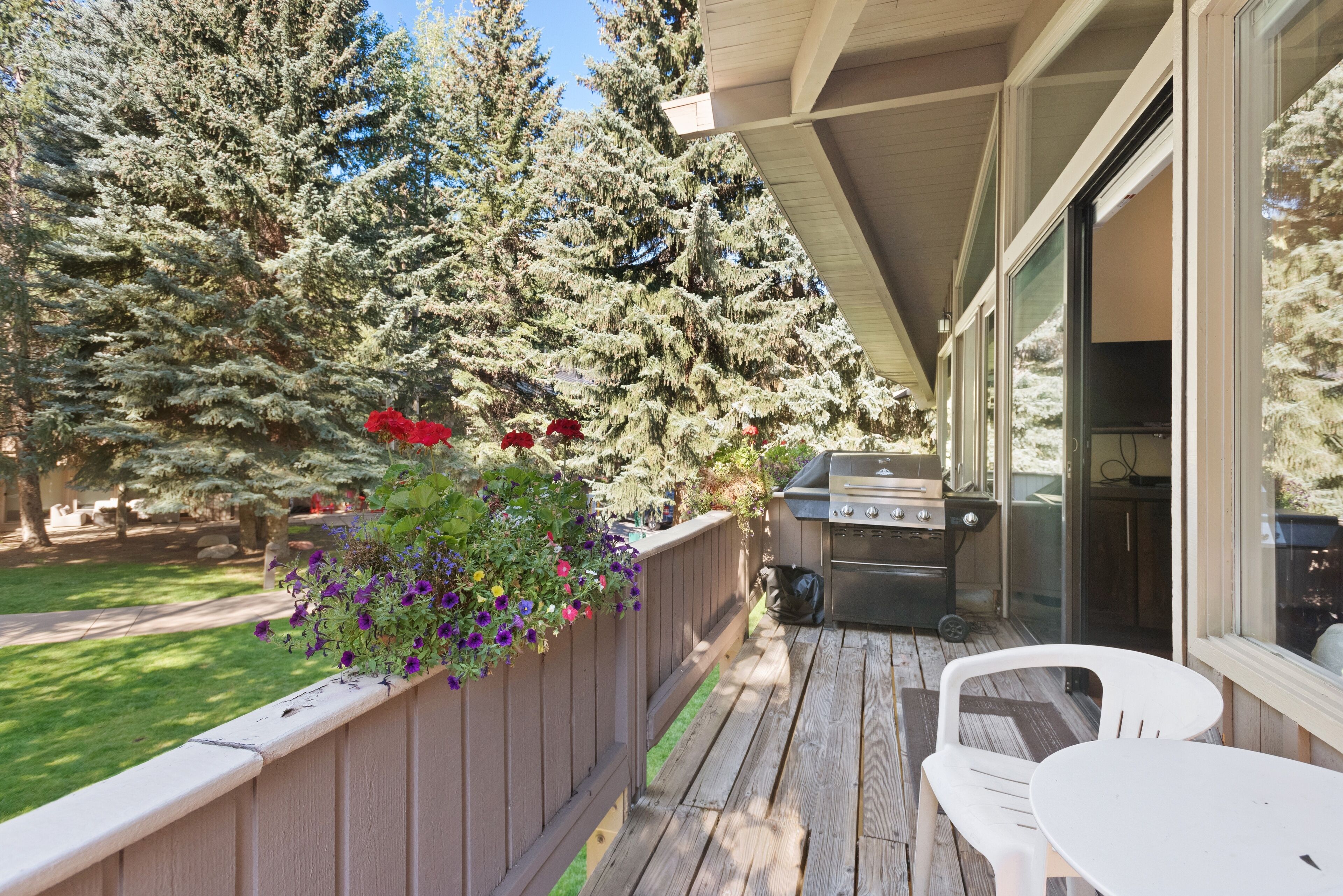 A wooden deck with a barbecue grill, white plastic chairs, and a flower planter with red flowers.
