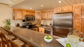 A kitchen with wooden cabinets, stainless steel appliances, a granite countertop, bar stools, and a pitcher of lemonade. - Comfortable & Modern 2 Bedroom Apartments - Right on the Slopes! (Aspen)