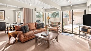 A spacious living room with a brown leather sofa, a wooden coffee table, and a television. There is a sliding glass door leading to a patio area. - Comfortable & Modern 2 Bedroom Apartments - Right on the Slopes! (Aspen)