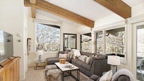 A modern living room with a sectional sofa, a coffee table, and a television on a wooden cabinet. Large windows provide a view of trees and a mountain. - Two Bedroom Apartments with One of a Kind Location on Slopes of Aspen Mountain! (Aspen)