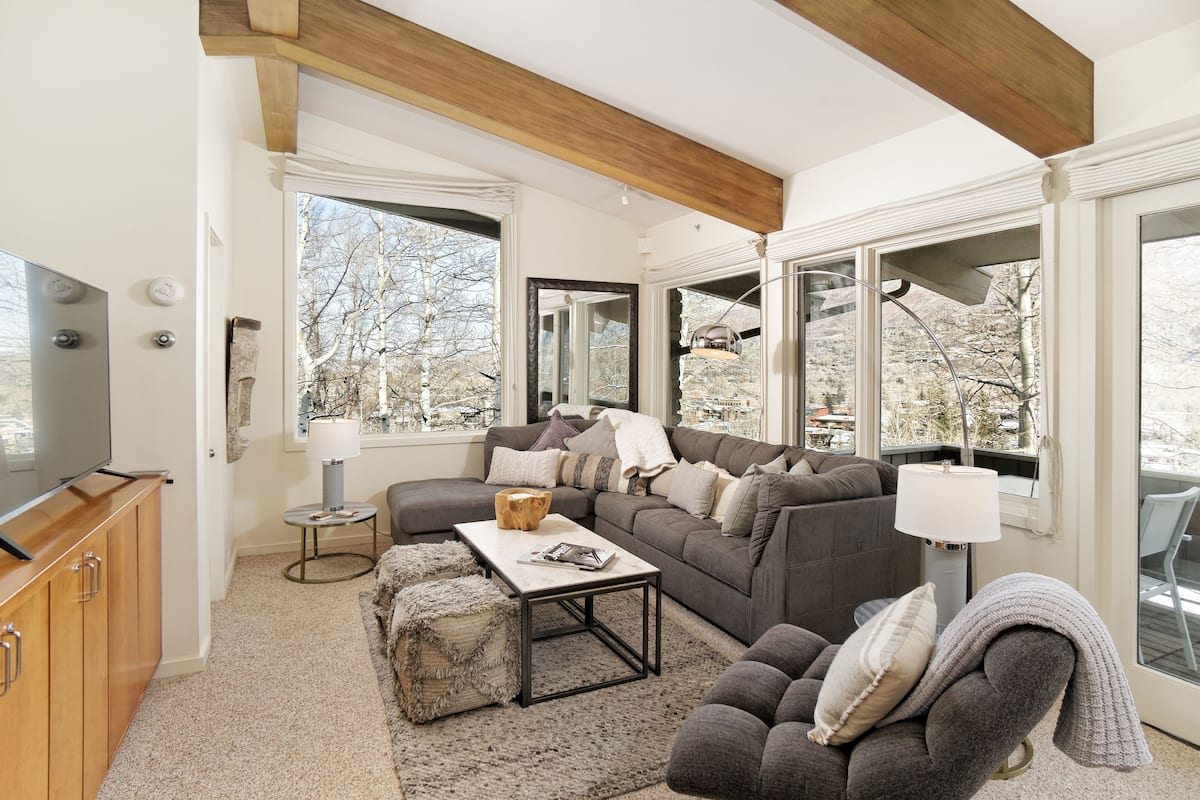 A modern living room with a sectional sofa, a coffee table, and a television on a wooden cabinet. Large windows provide a view of trees and a mountain.