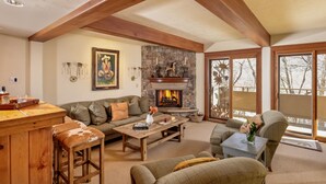 A cozy living room with a stone fireplace, wooden beams, a sofa, a coffee table, and a view through large windows. - Deluxe 3 & 4 Bedroom Aspen Apartments Next to Silver Queen Gondola (Aspen)
