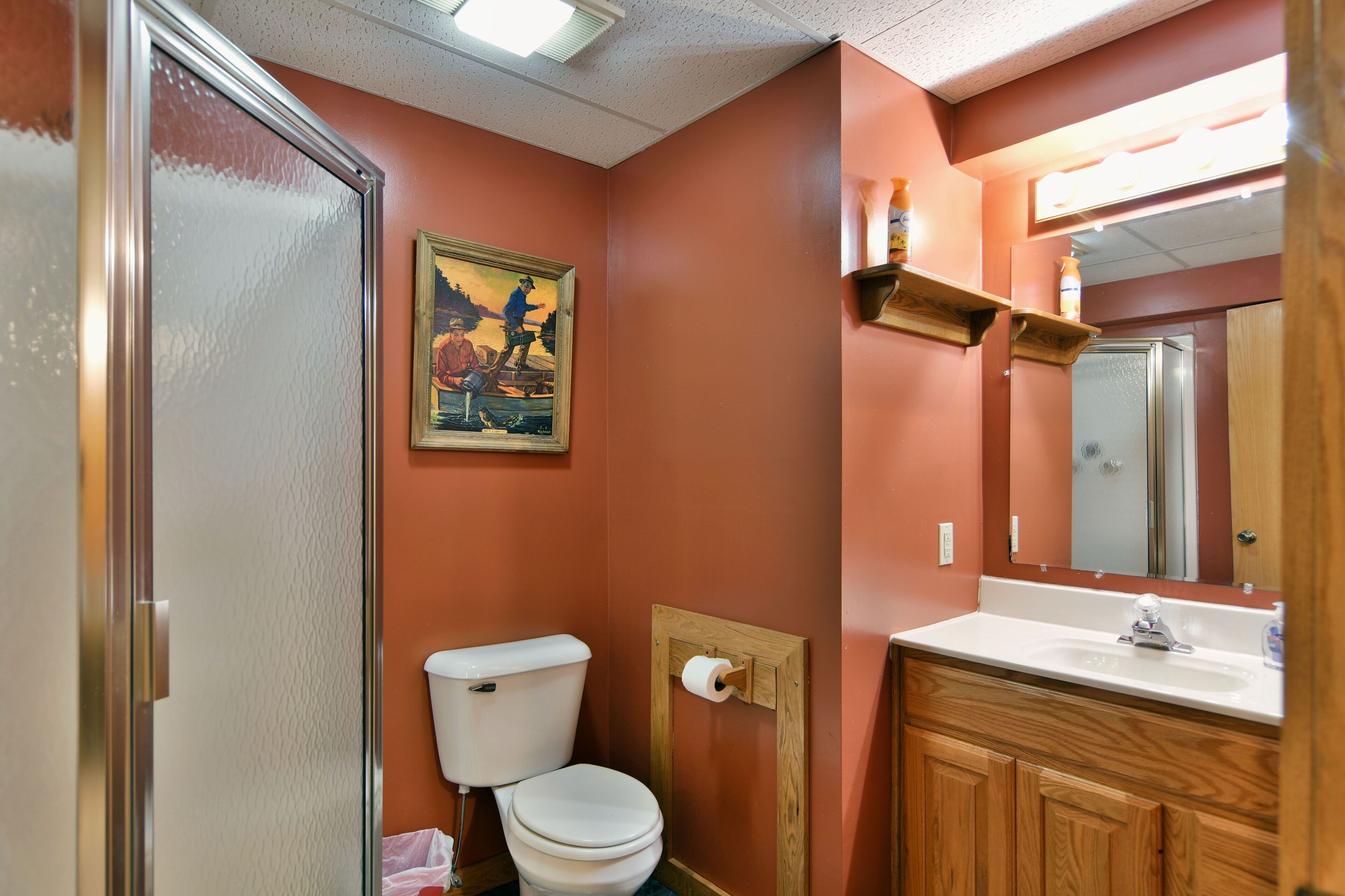 A bathroom with a toilet, sink, wooden cabinets, a mirror, and a painting on the wall.