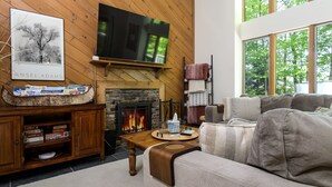 A cozy living room with a fireplace, a flat-screen TV, and a large window offering a view of trees. - Okemo Westwood Condo 3 Bedroom Condo by RedAwning (Ludlow)