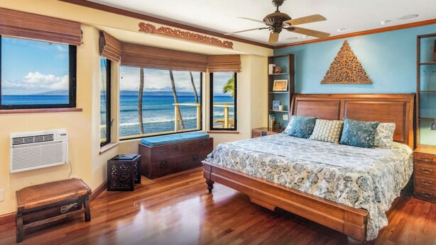 Een slaapkamer met een groot bed, houten vloer, een plafondventilator en uitzicht op de oceaan door grote ramen.