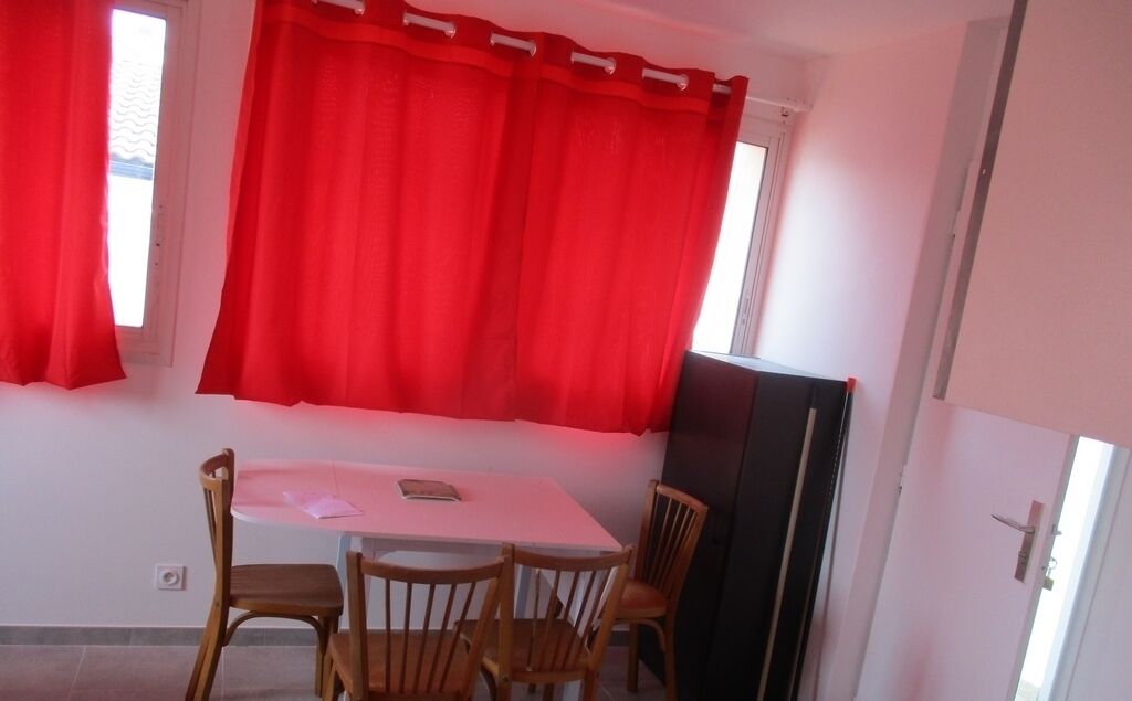 A room with a dining table, four wooden chairs, a black cabinet, and red curtains.