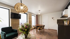 A modern living room with a round table, two green armchairs, a decorative vase with flowers, a large woven light fixture, and a kitchen area with a dark countertop and a built-in oven. - My Nest Inn Marais - 5 pers- 53m² - Heart of Paris (Paris)