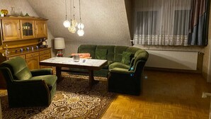 Un salón acogedor con un sofá verde, dos sillones, una mesa de centro de madera y una alfombra estampada. Contra la pared, se encuentra un aparador de madera con puertas de cristal, y se observa un radiador.