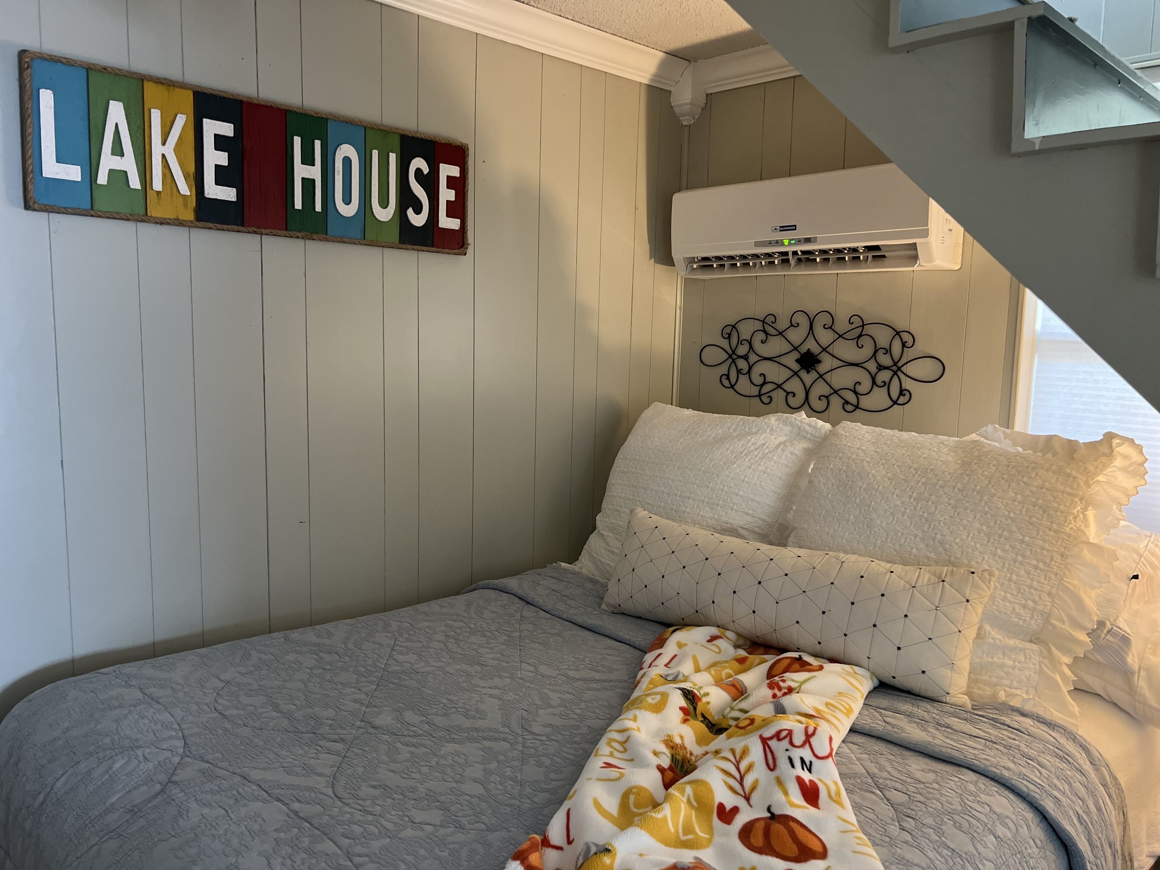 Une chambre à coucher comprenant un lit, des oreillers, une couverture, un climatiseur fixé au mur et un panneau décoratif sur lequel est inscrit « MAISON AU BORD DU LAC ».