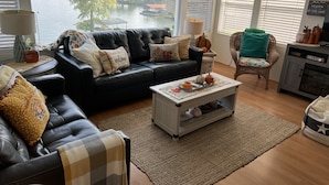 Un salon avec un canapé en cuir noir, une table basse et un fauteuil en osier. De grandes fenêtres offrent une vue sur un lac.