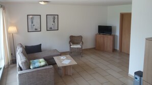 Una sala de estar con un sofá, una mesita, una silla de mimbre, un televisor sobre un mueble de madera y dos cuadros enmarcados en la pared.