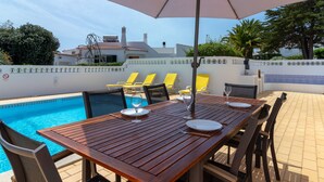Mesa de madera dispuesta para comer con vista a una piscina, sillones de descanso amarillos y un paraguas grande.