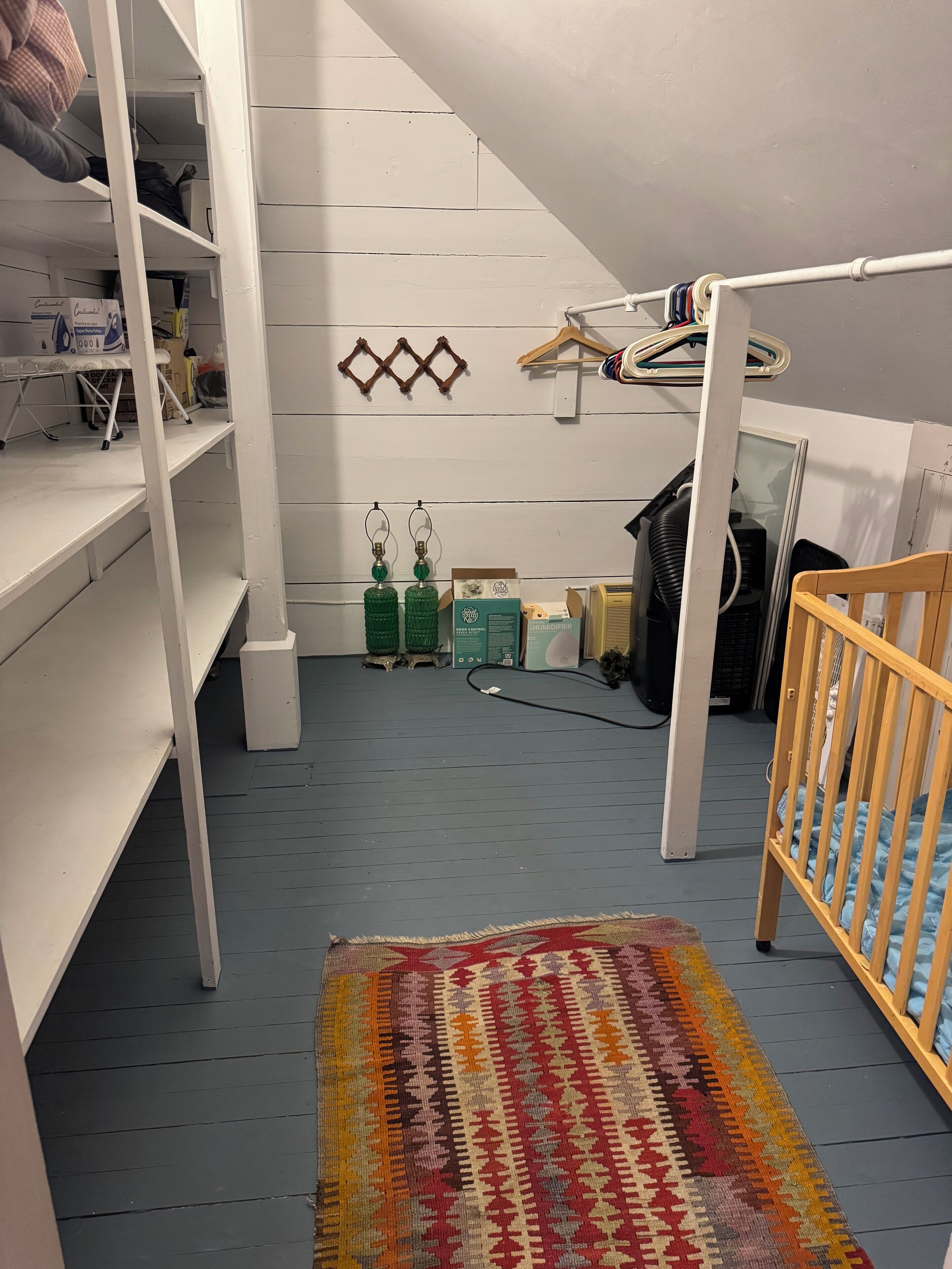 Una habitación con una cama litera, una cuna pequeña, un tapete colorido, un estante blanco y una percha con ropa.