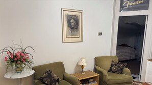 Una sala de estar con dos sillones verdes, una mesita auxiliar blanca con una lámpara, un cuadro enmarcado en la pared y un jarrón floral sobre una mesita.