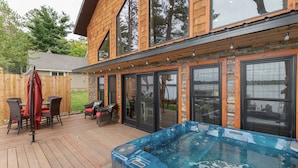 Une terrasse en bois avec un jacuzzi, du mobilier d’extérieur et une grande fenêtre donnant sur une cour.