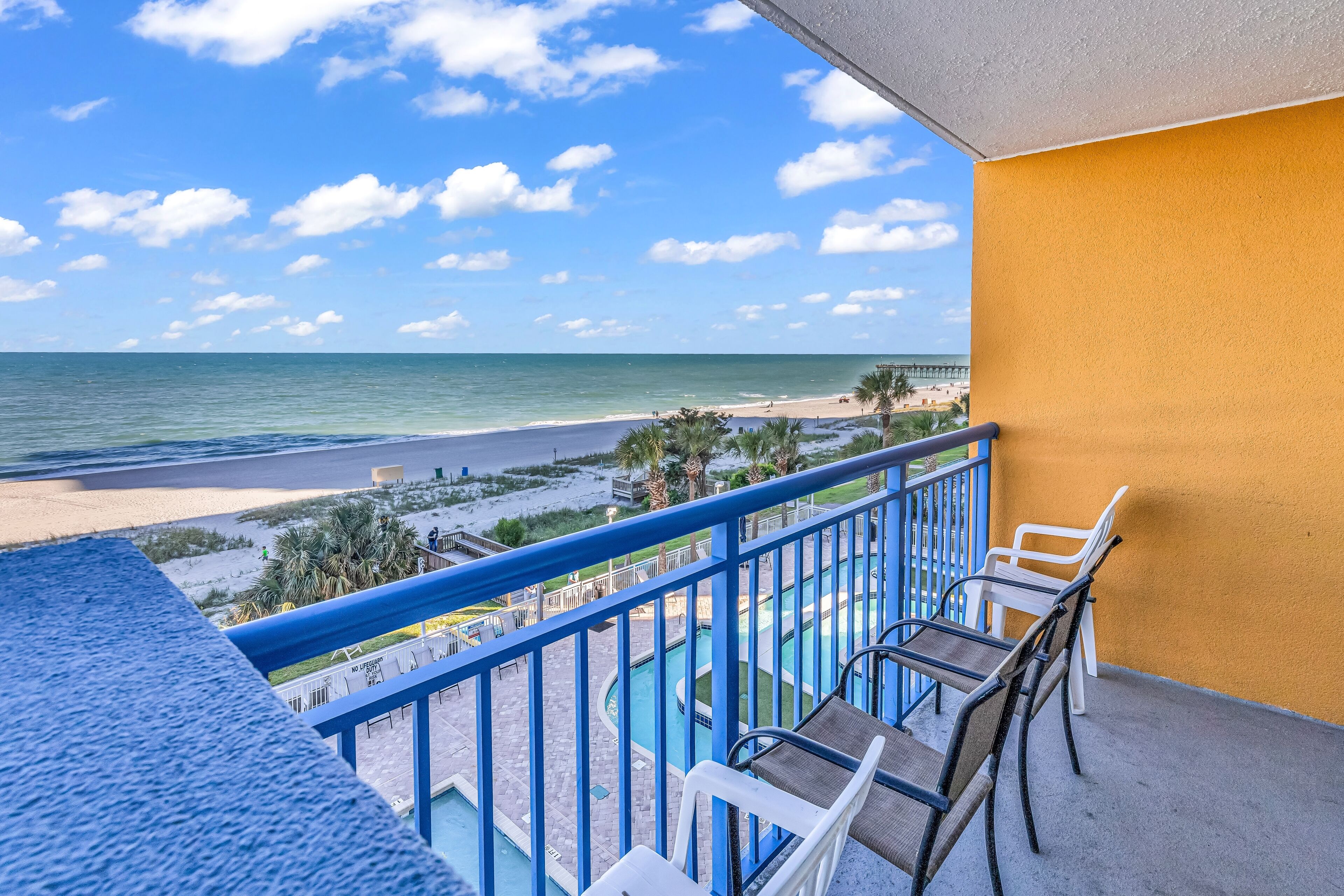 A balcony with a view of the beach and ocean, featuring a pool area with chairs and a railing.