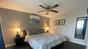 Une chambre à coucher comprenant un lit, deux lampes de chevet, un ventilateur de plafond, un appareil de climatisation et des tableaux encadrés accrochés au mur.