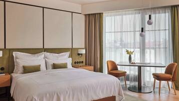 A modern hotel room with a large bed, a round table with chairs, a small desk, and a large window with curtains.