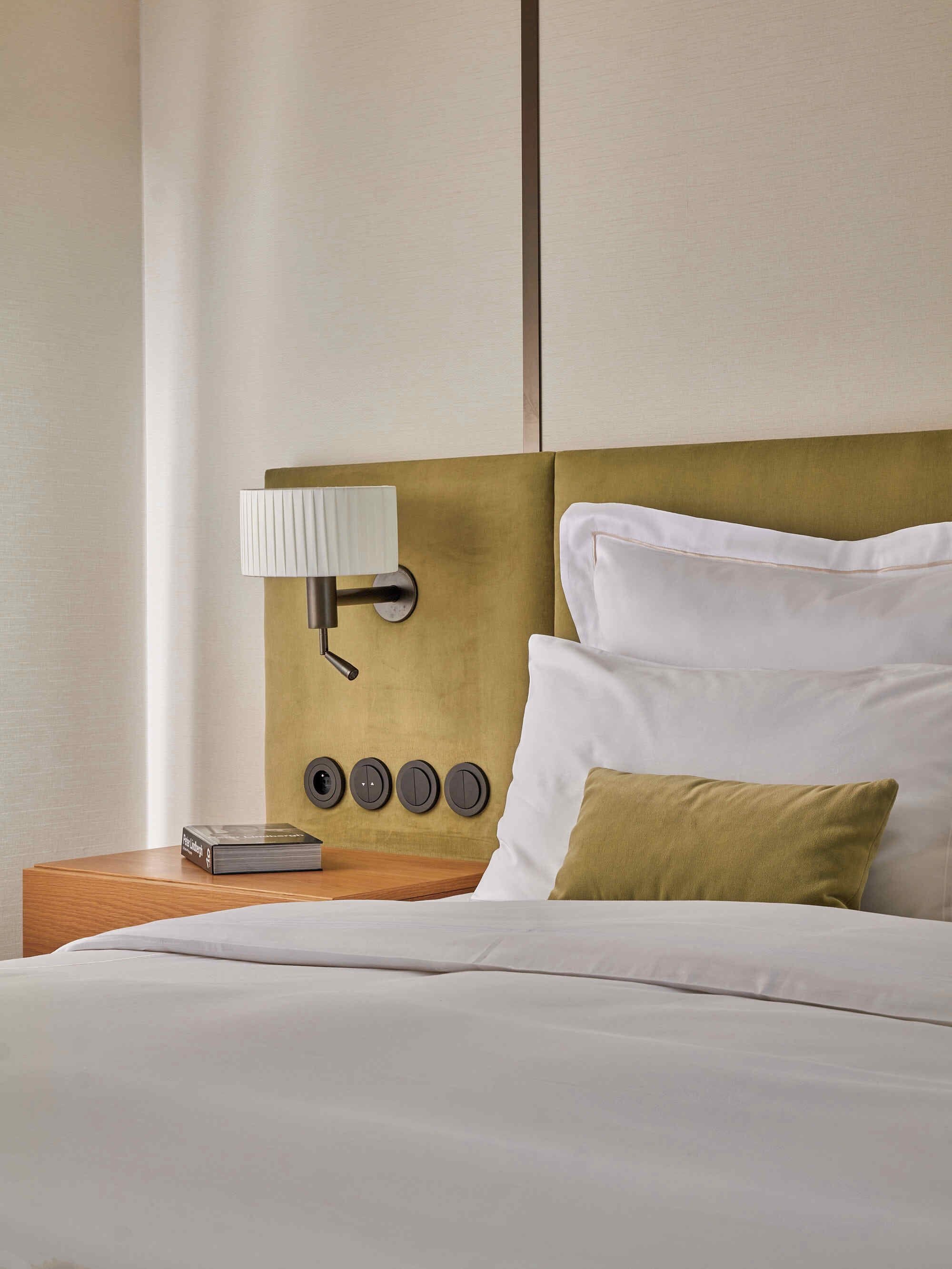 A neatly made bed with white linens, a gold-toned headboard, a bedside lamp, and a book on the nightstand.