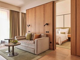 A modern hotel room with a sofa, a small round table with fruit, a bed with white bedding, and wooden paneling.