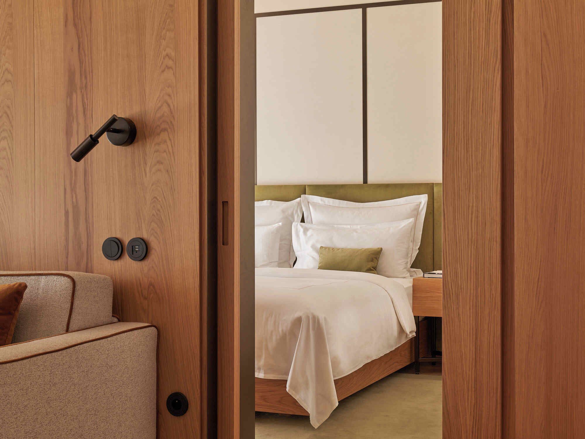 Una habitación de hotel con una puerta de madera, una cama con ropa blanca y un sofá color beige.
