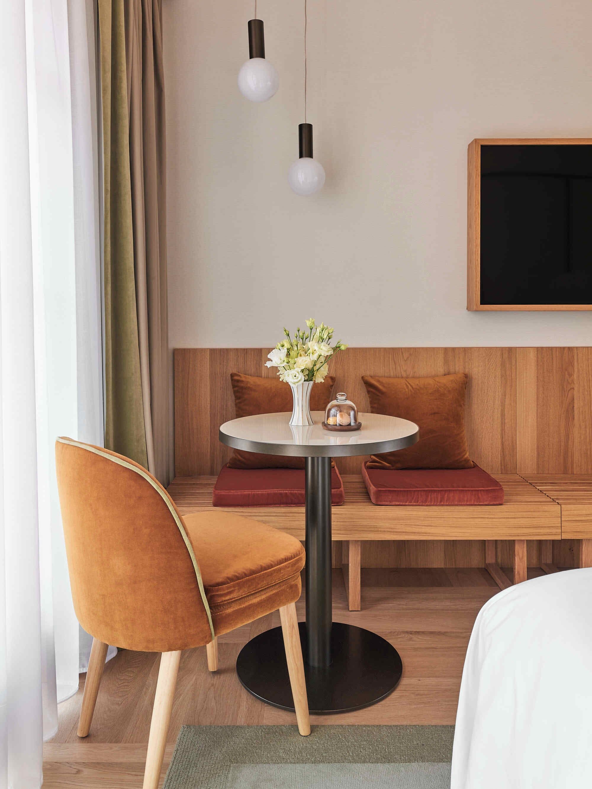 A hotel room with a wooden bench, a round table with a vase of flowers, a bed, and a TV mounted on the wall.