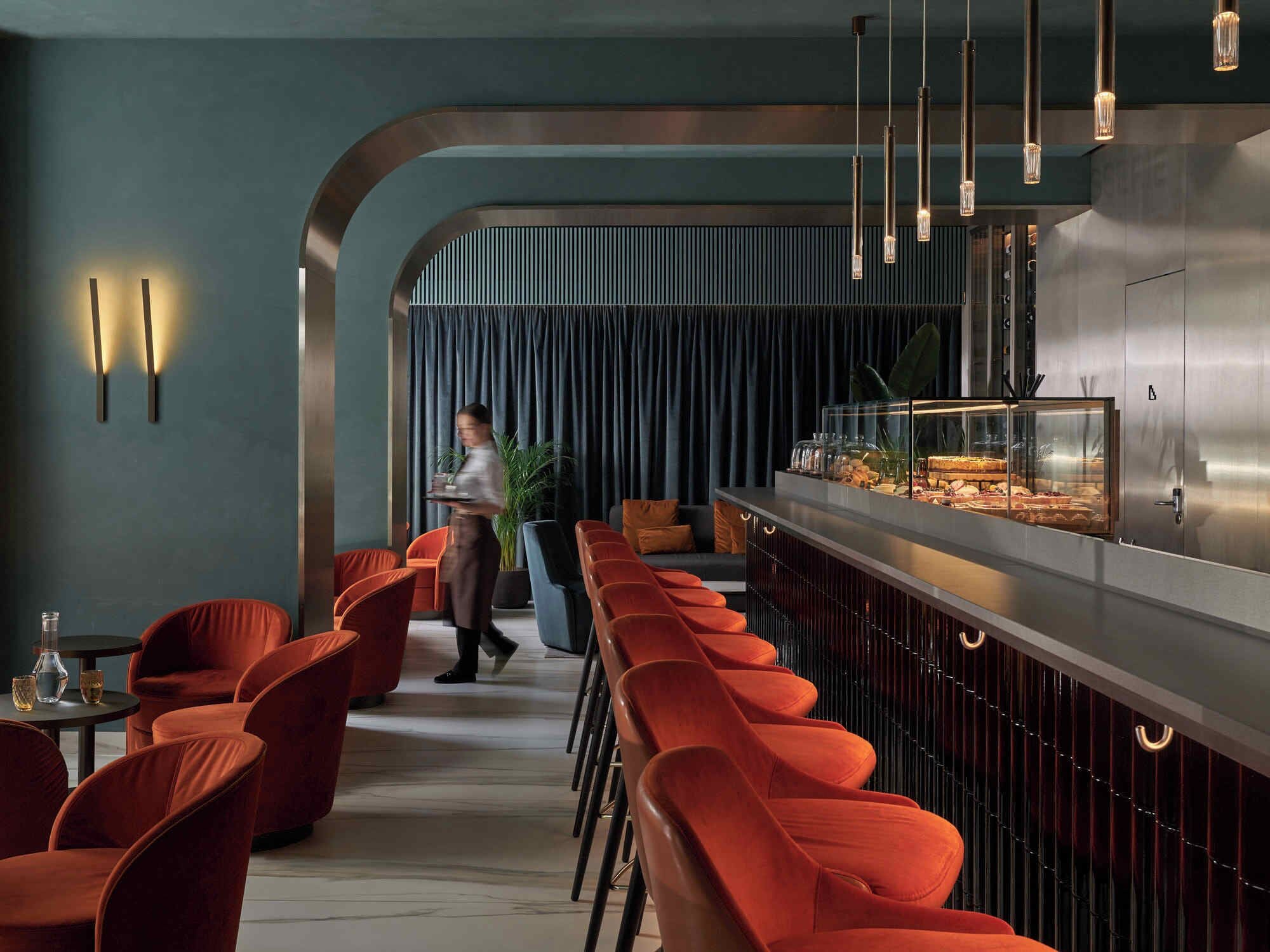 Un espace bar moderne avec des chaises à revêtement rouge, un comptoir en acier inoxydable et des luminaires suspendus.
