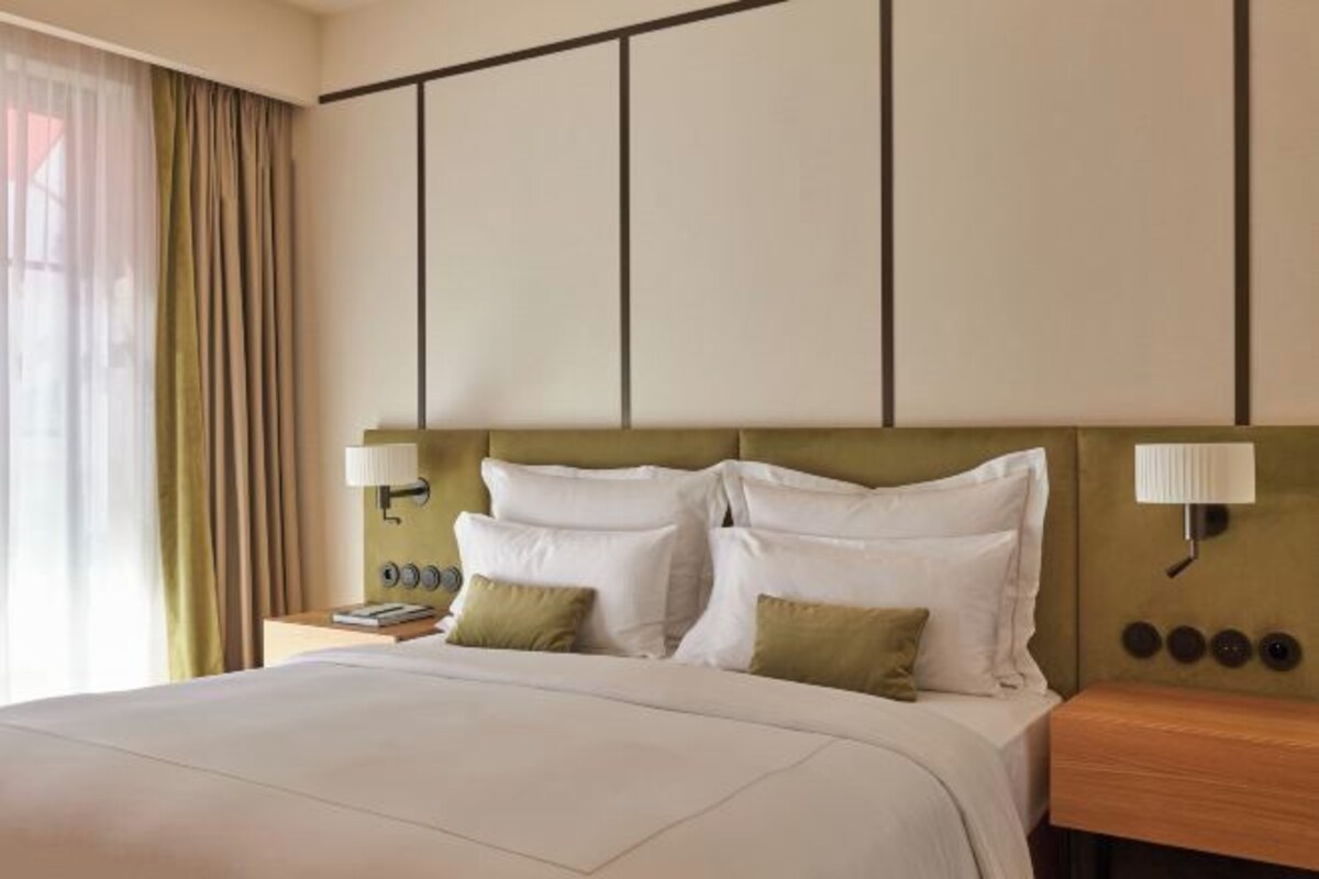 A hotel room with a large bed, two bedside tables, a headboard with a light fixture, and a window with curtains.