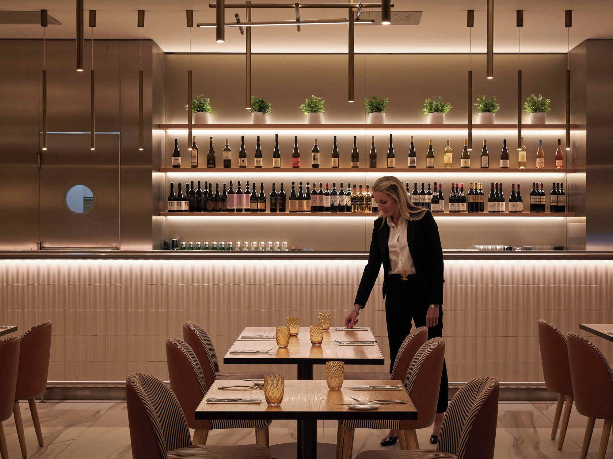 A modern bar area with neatly arranged bottles and a person attending to a table.