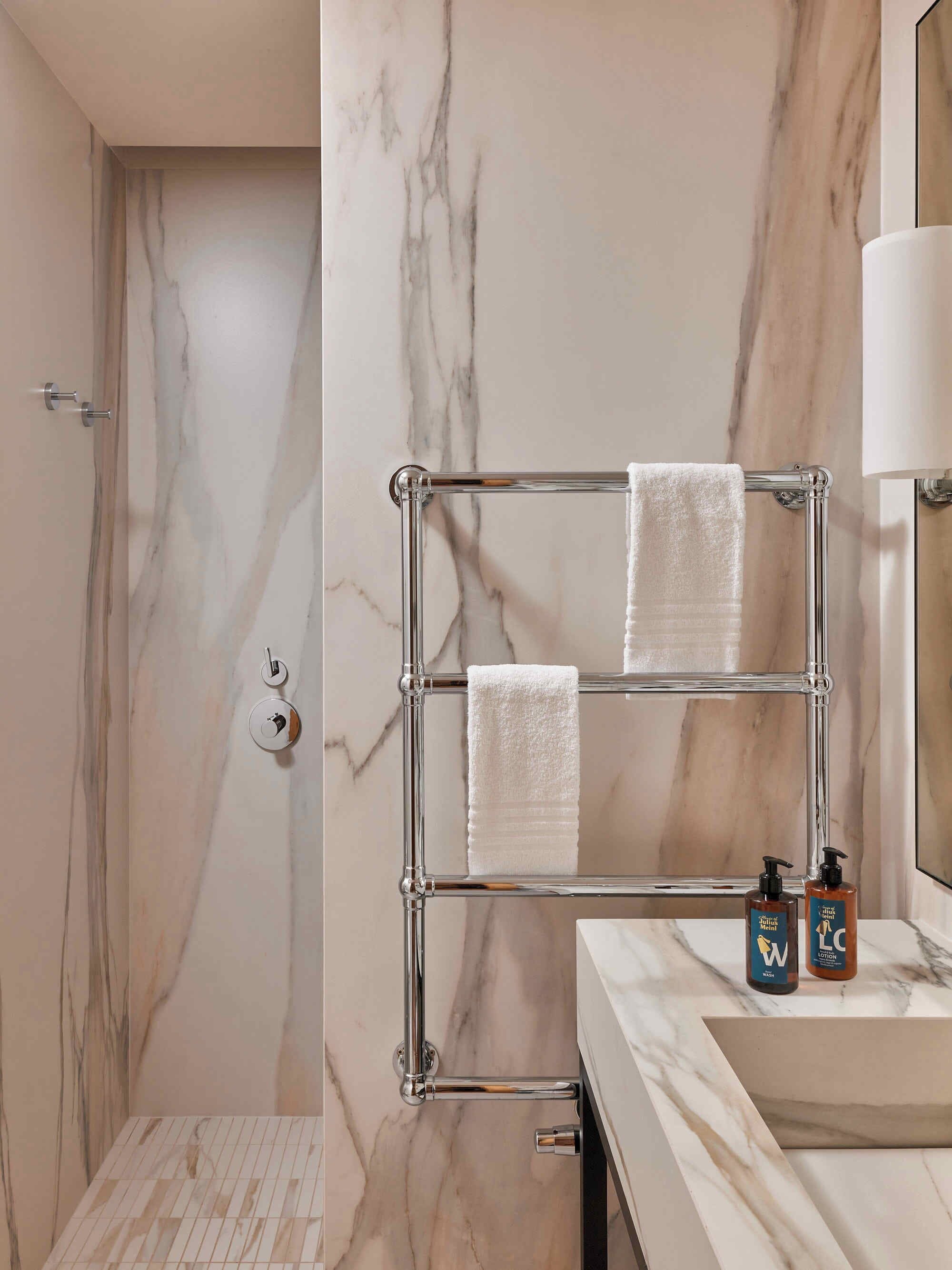 A bathroom with a marble wall, a glass shower enclosure, and a towel rack with two towels. There are two bottles of lotion on the sink.