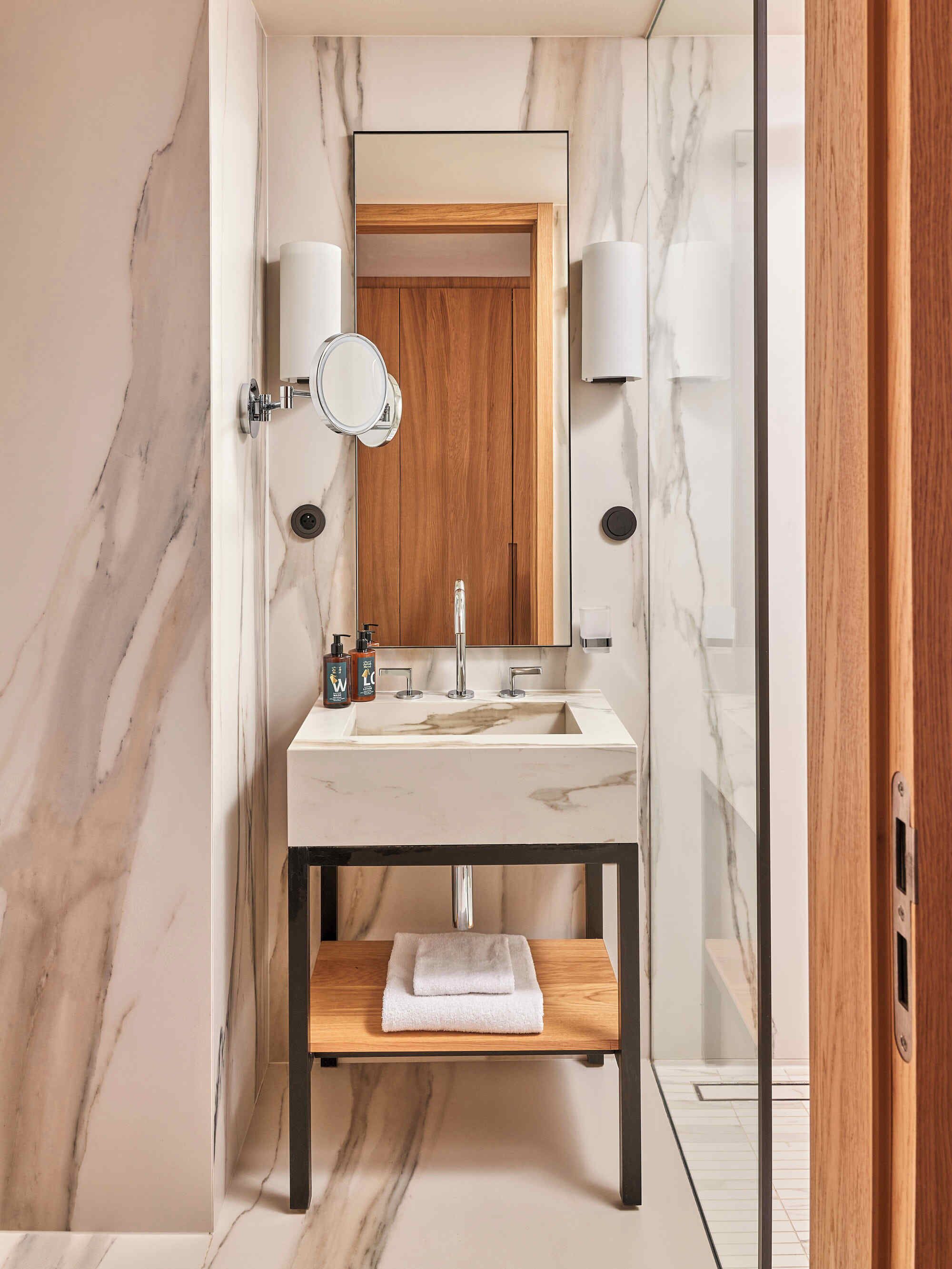 A modern bathroom with a marble wall, a wooden door, a sink with a mirror, and two wall-mounted lights.