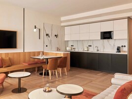 Uma cozinha moderna com ilha central, armários escuros e micro-ondas. Há uma área de refeições com mesa e cadeiras, e um sofá na sala de estar.