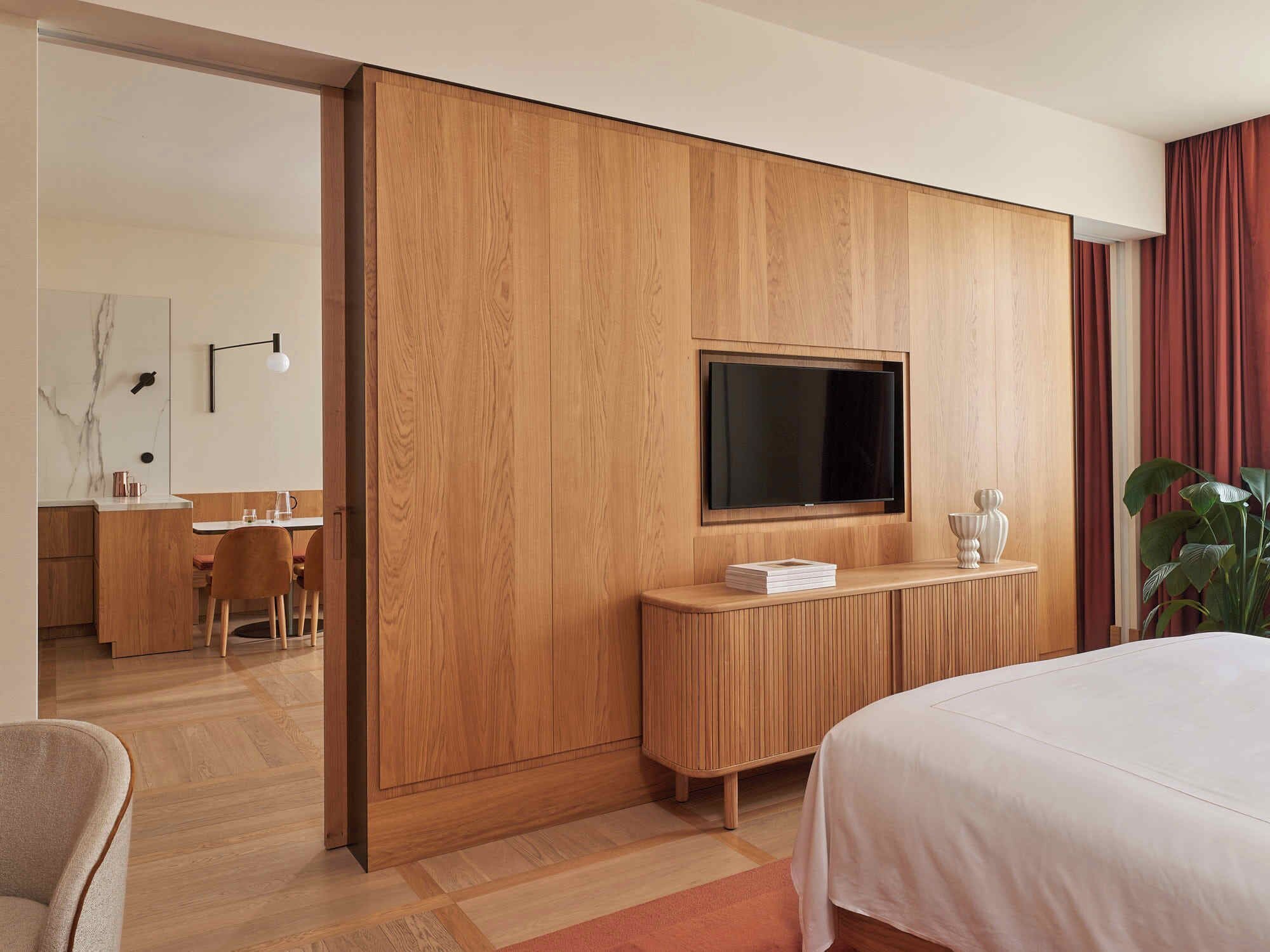 Una habitación de hotel moderna con una pared de paneles de madera, un televisor de pantalla plana montado sobre un mueble de madera, una cama con colcha blanca y una zona de comedor visible a través de una puerta abierta.
