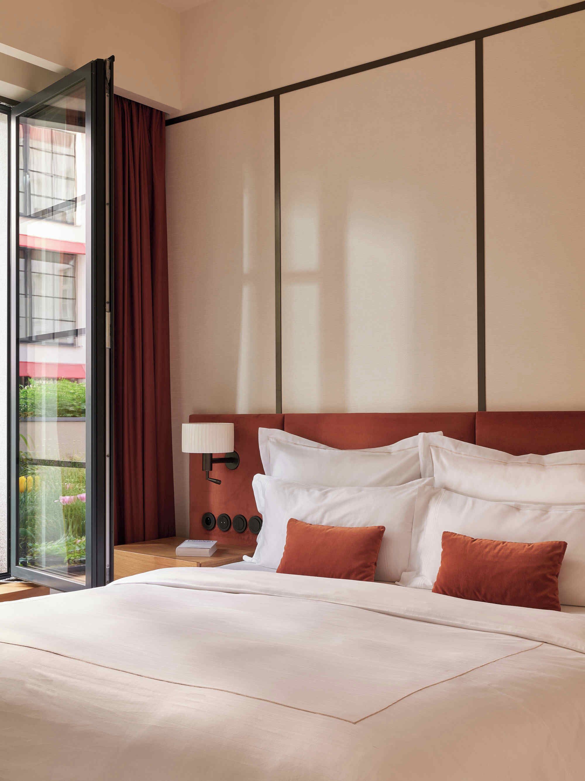 A hotel room with a large bed, a nightstand with a lamp, and a view of a building through a window with curtains.