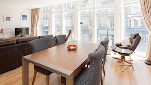 Uma sala de estar moderna com uma mesa de jantar de madeira, cadeiras estofadas cinzentas, um sofá de couro e uma mesa de centro de vidro.