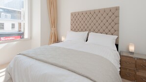 Uma cama bem arrumada com cabeceira estofada, mesa de cabeceira com abajur e janela com cortinas.