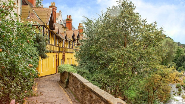 Uma rua de calçada de pedra leva a um edifício amarelo, com uma parede de pedra de um lado e vegetação exuberante do outro.