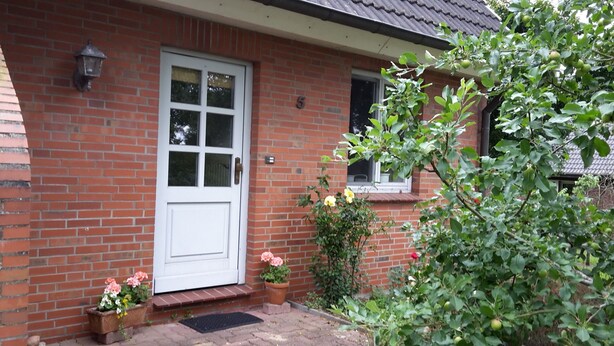 Ein Ziegelhaus mit weißer Tür, Hausnummer 5, und einem kleinen Garten mit Blumentöpfen und einem Baum.