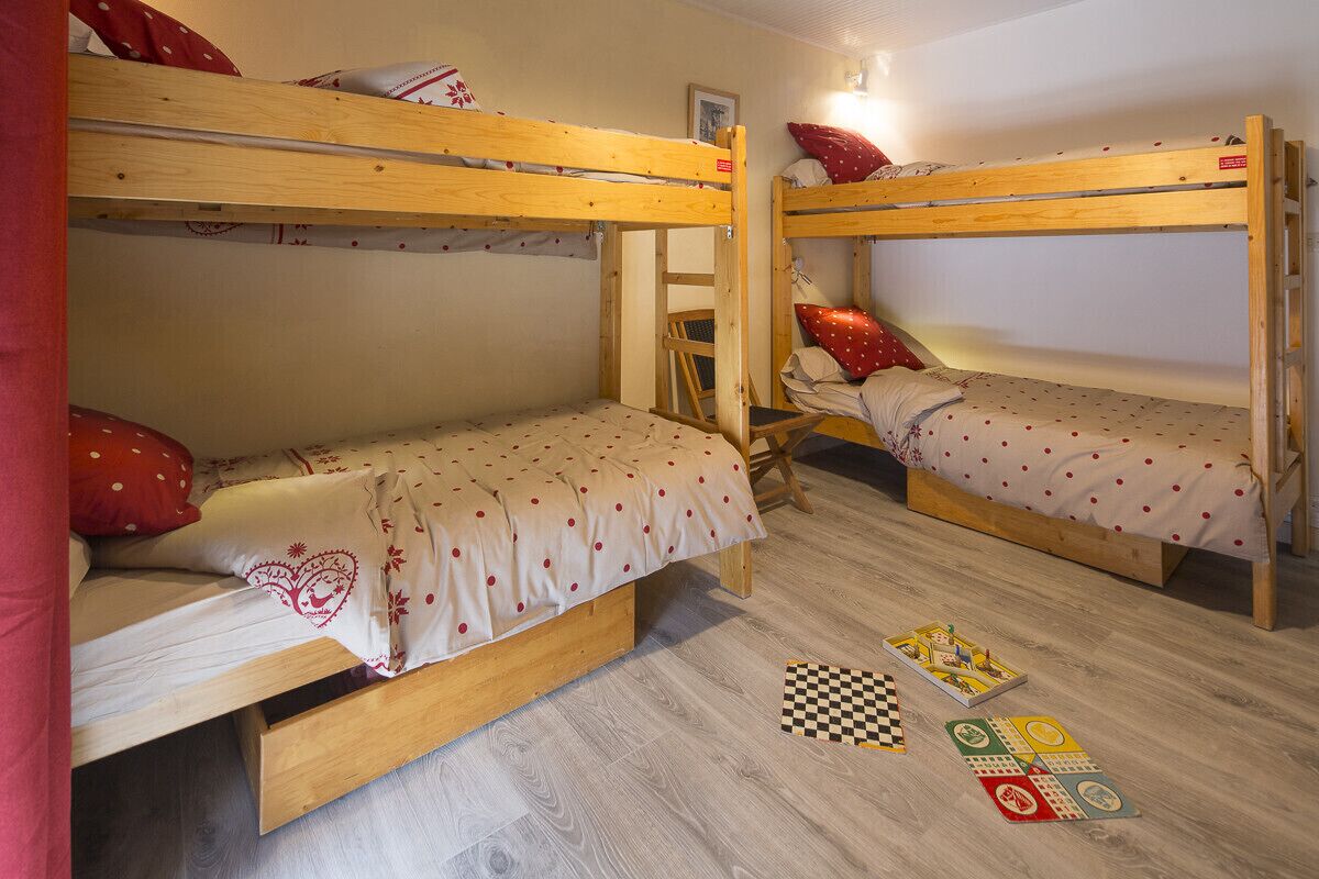 A room with two bunk beds, each with a wooden frame and a mattress with a polka dot cover. There is a chair in the center of the room and a checkered board game on the floor.