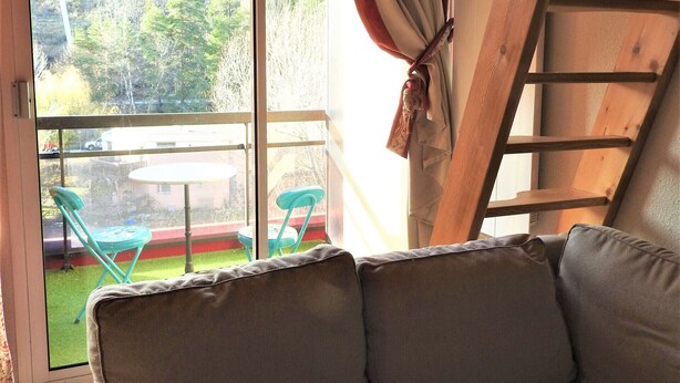 Een kamer met een bank, uitzicht op een groen gazon en bomen, en een houten ladder.