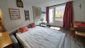 A bedroom with a bed, a desk, a chair, a window with curtains, a clock, and a cross on the wall.